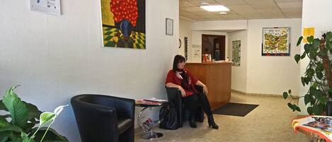 Una zona de recepción con una recepcionista sentada, un sofá negro, una mesita con revistas y una planta en maceta.