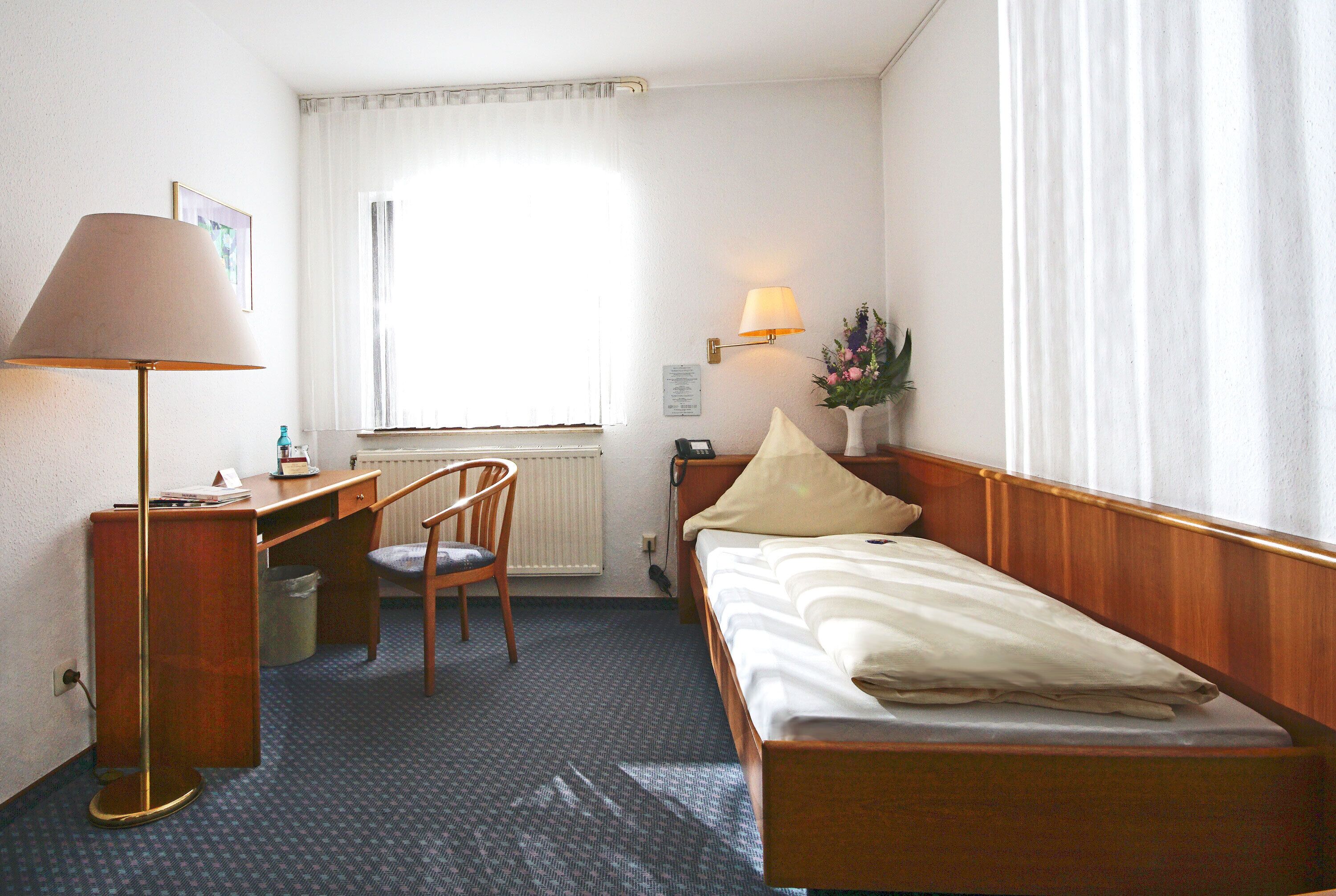Een hotelkamer met een enkel bed, een bureau met stoel, een lamp, een radiator, een raam met gordijnen en een wandlamp.