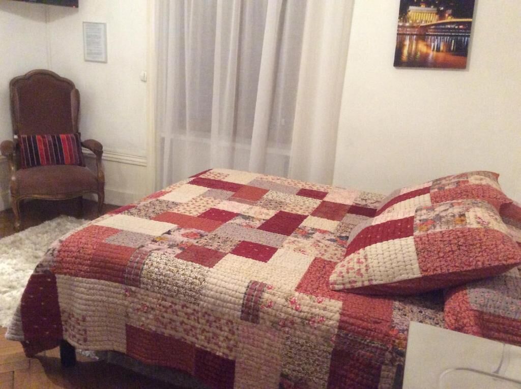 Un dormitorio con una colcha acolchada, una silla, una ventana con cortinas y un cuadro en la pared.