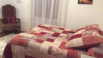 Un dormitorio con una colcha acolchada, una silla, una ventana con cortinas y un cuadro en la pared.