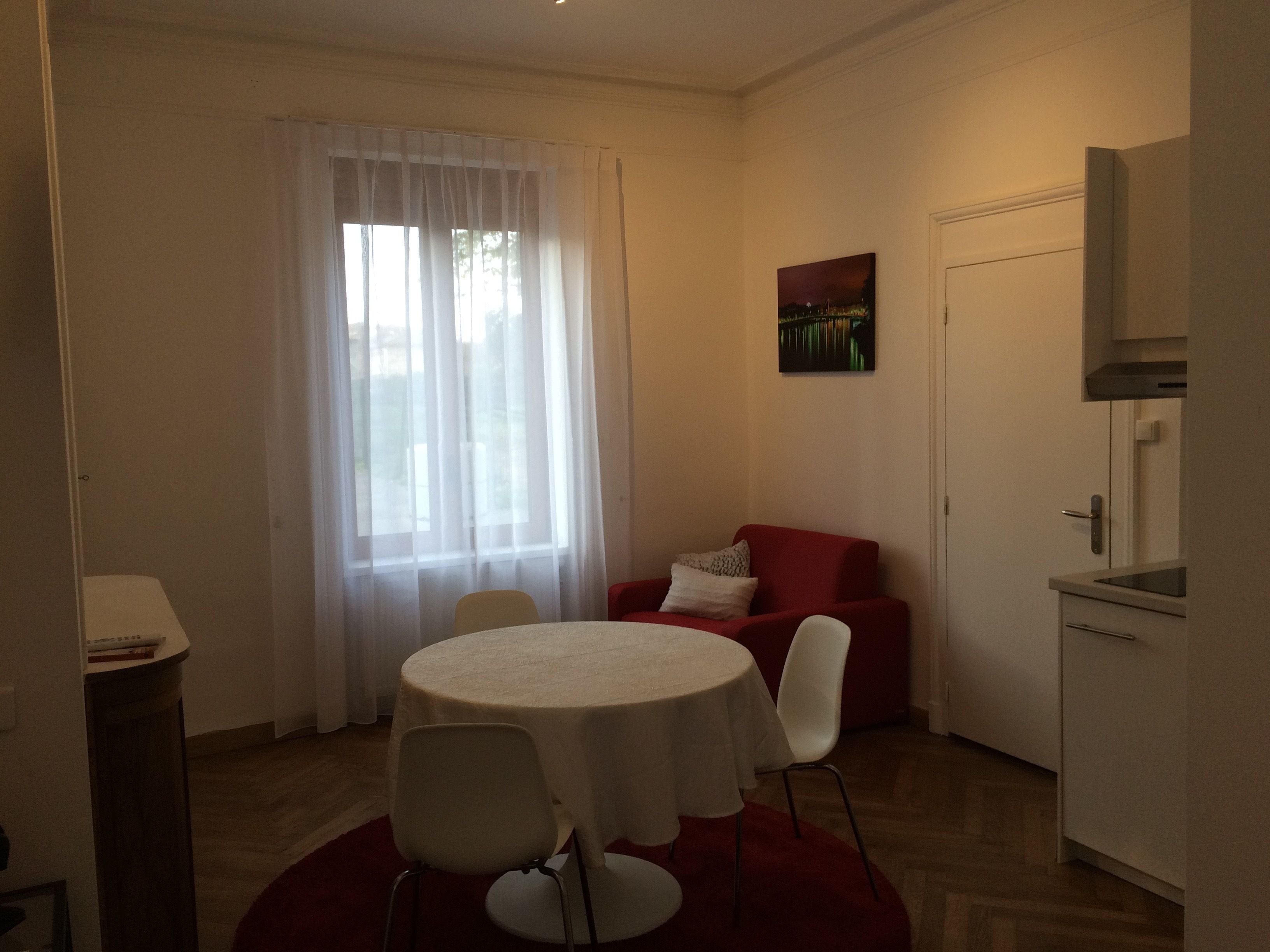 A compact living space with a dining area, a red sofa, a kitchen area, and a window with sheer curtains.