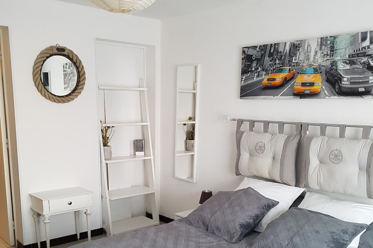 Un dormitorio con una cama, una mesita de noche blanca, un tocador blanco, un espejo y un estante en la pared.