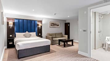 A hotel room with a bed, a sofa, a small table, a shower area, and a wall-mounted light fixture.