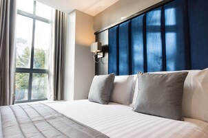 Una habitación de hotel moderna con una cama grande, una ventana con cortinas, una lámpara de pared y un cabecero azul.