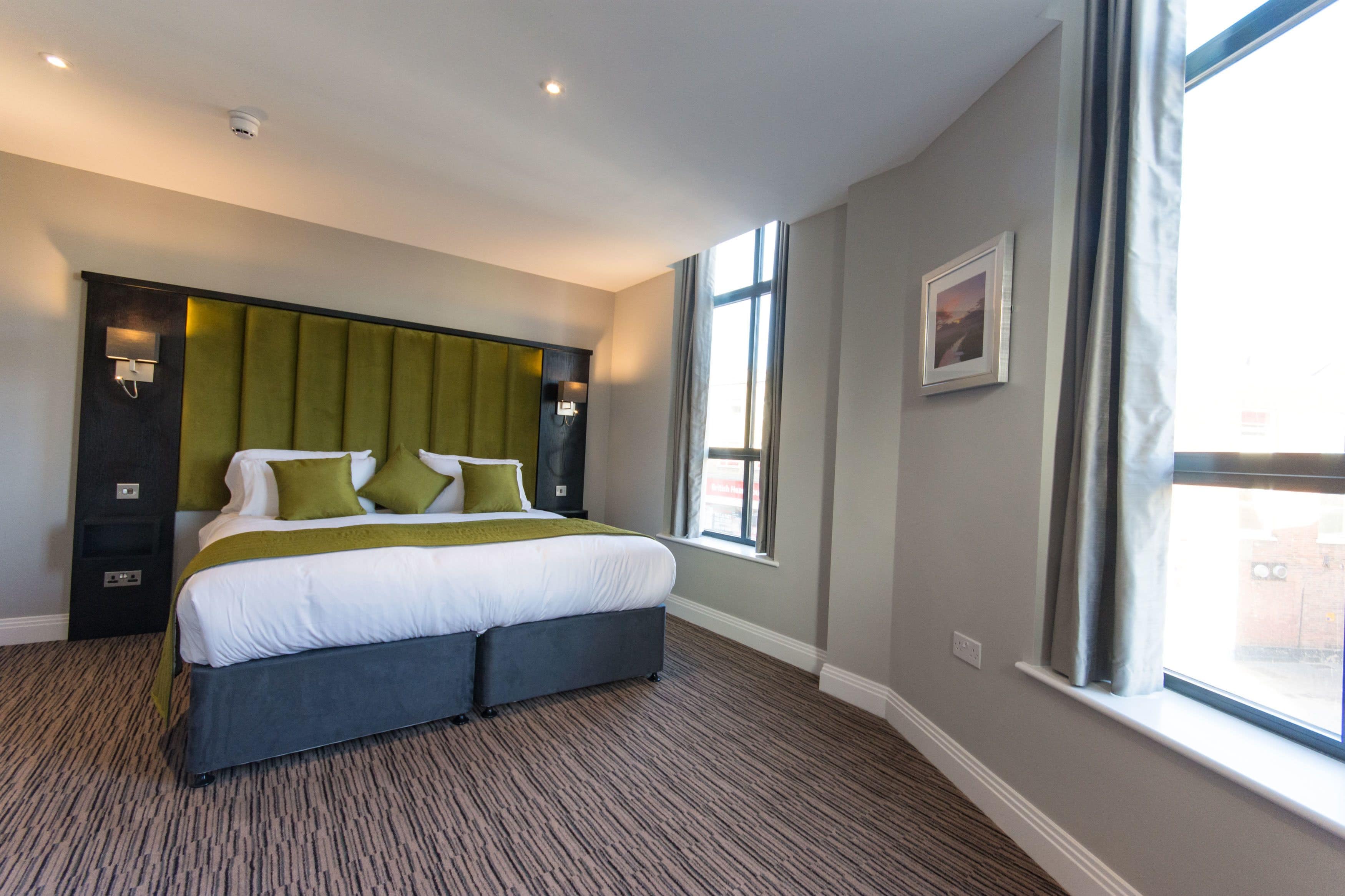 A modern hotel room with a large bed, a headboard with built-in lighting, a window with curtains, and a carpeted floor.