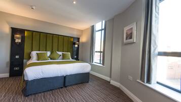 A modern hotel room with a large bed, a headboard with built-in lighting, a window with curtains, and a carpeted floor.