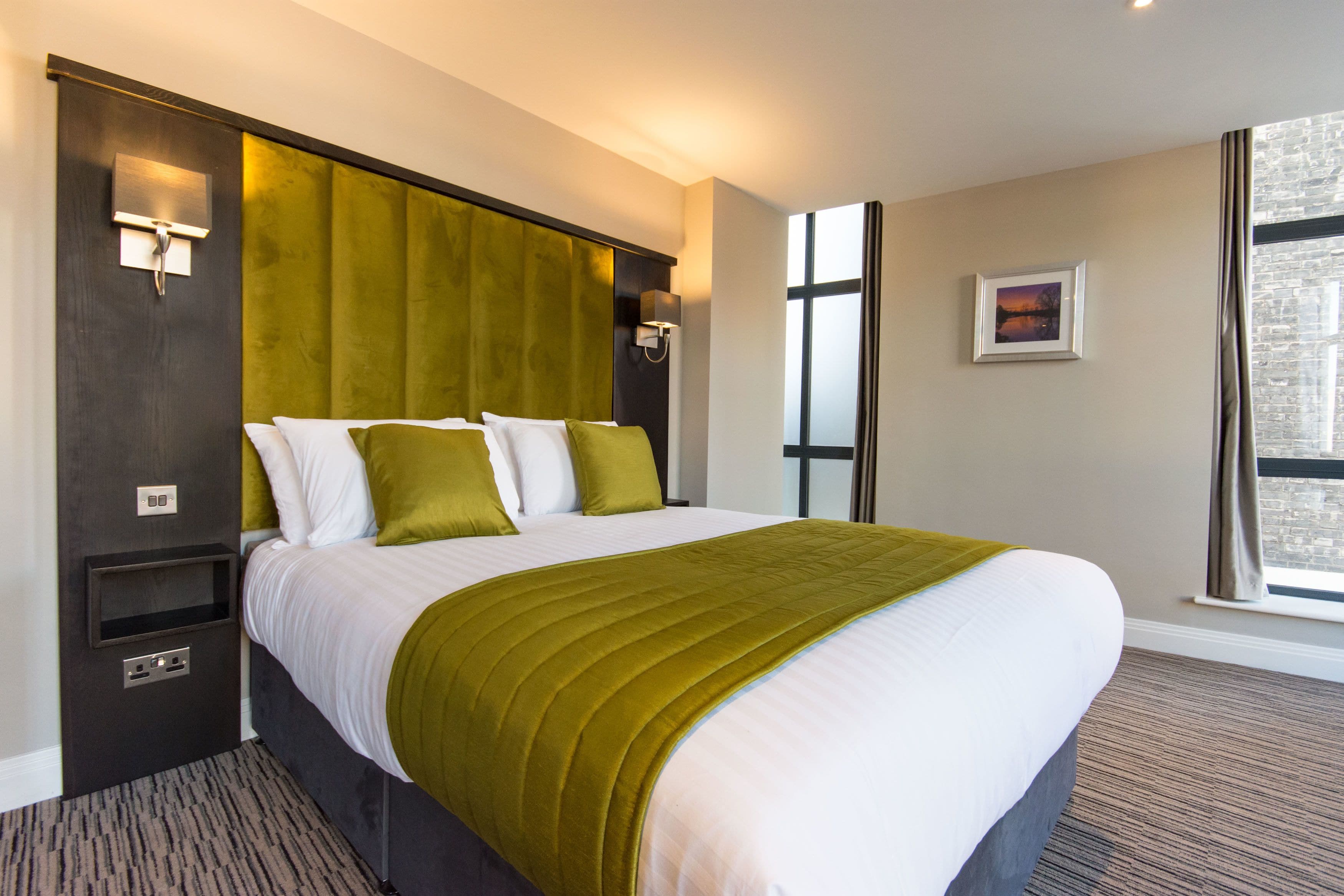 A hotel room with a large bed, a headboard with a light fixture, a bedside table, and a window with a view.