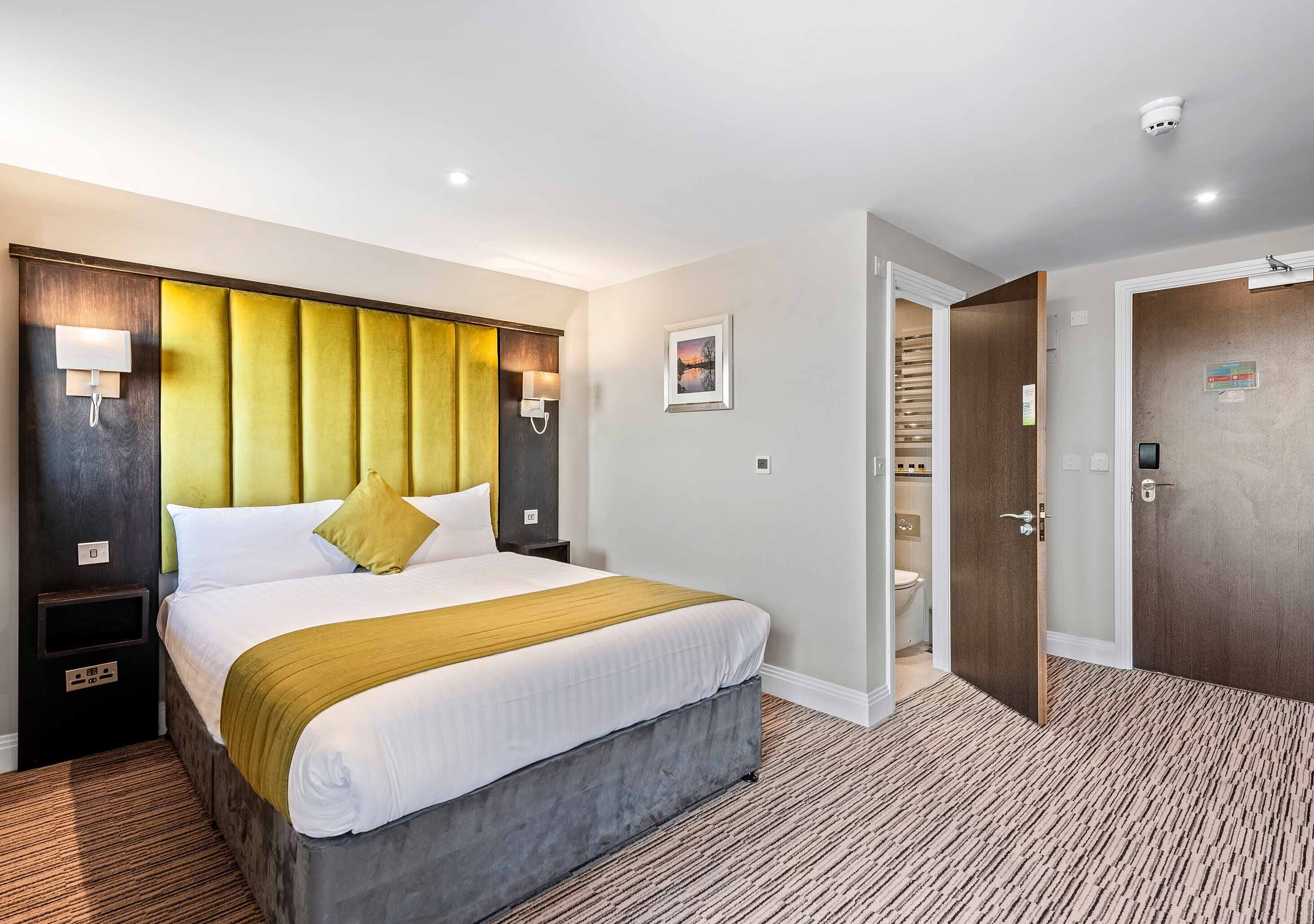 A hotel room with a bed, bedside tables, a nightstand, a wall-mounted lamp, a picture, and a door leading to a bathroom.