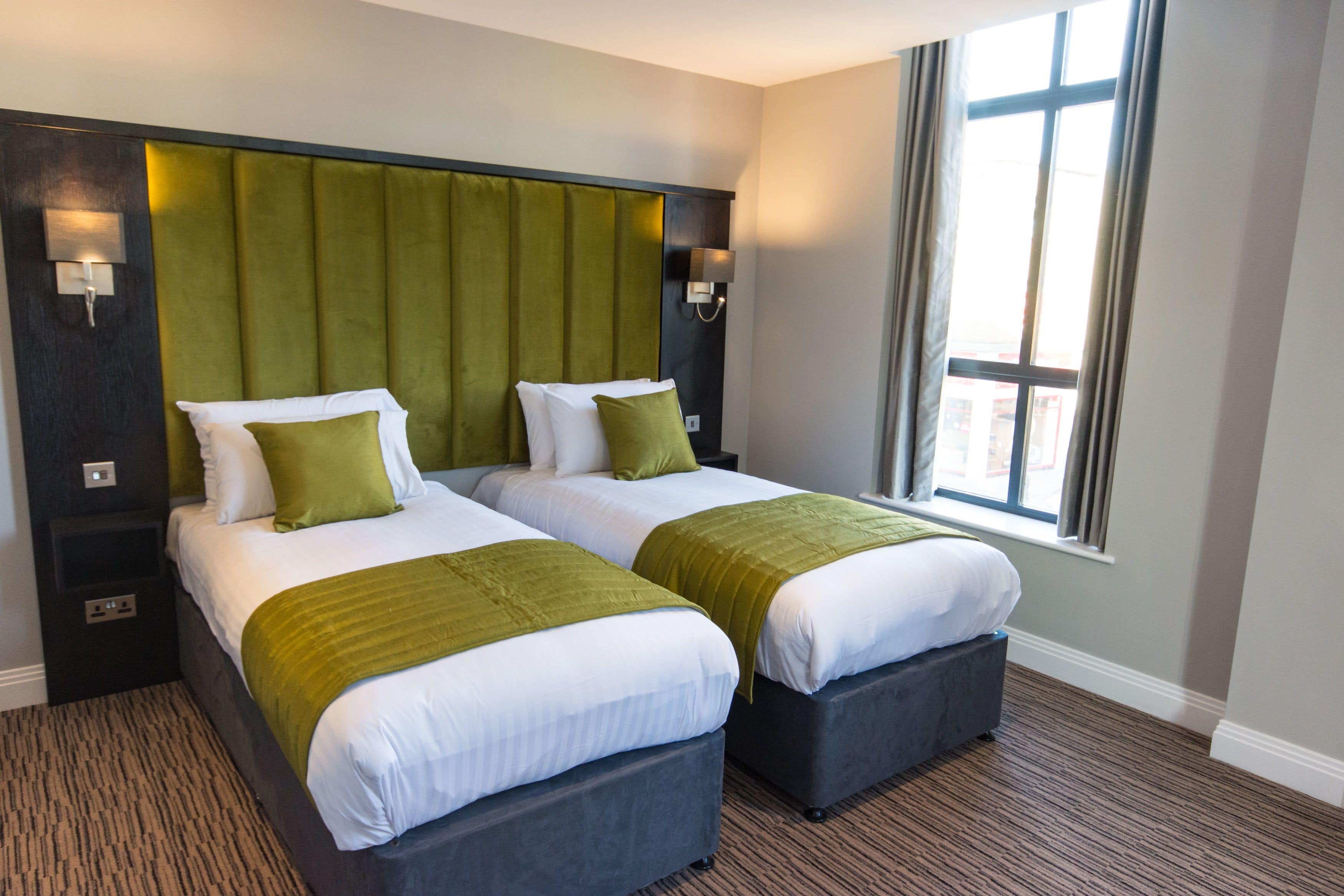 Une chambre d’hôtel avec deux lits, chacun doté d’une tête de lit verte et de linge de lit blanc. Des lampes murales sont fixées de chaque côté de la tête de lit, et une fenêtre avec des rideaux se trouve sur le côté droit.