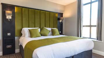 Ein Hotelzimmer mit einem großen Bett, grünen Kissen, einer grünen Tagesdecke, einem Kopfteil mit integrierter Beleuchtung und einem Fenster mit Vorhängen.