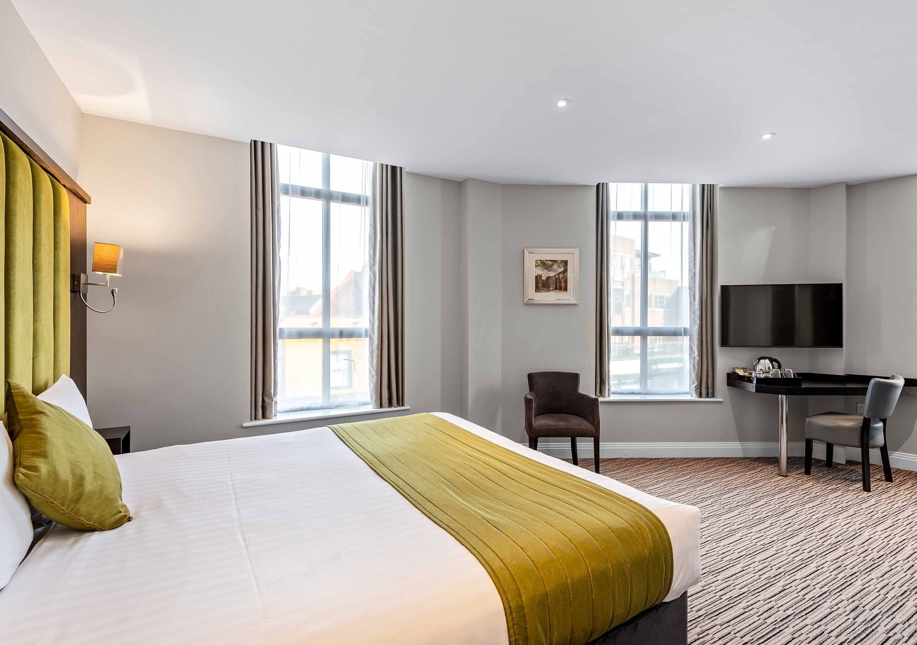 A hotel room with a large bed, a desk, and a chair. There are two windows with curtains, a flat-screen TV mounted on the wall, and a framed picture on the wall.