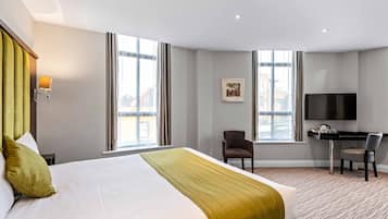Habitación de hotel con una cama grande, un escritorio y una silla. Hay dos ventanas con cortinas, un televisor de pantalla plana montado en la pared y un cuadro enmarcado en la pared.