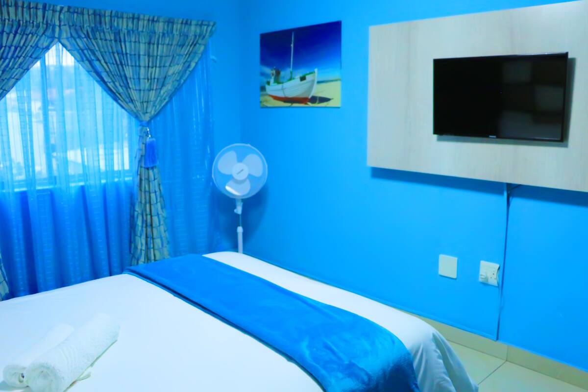 A bedroom with a bed, a TV mounted on the wall, a fan, and a painting of a boat.