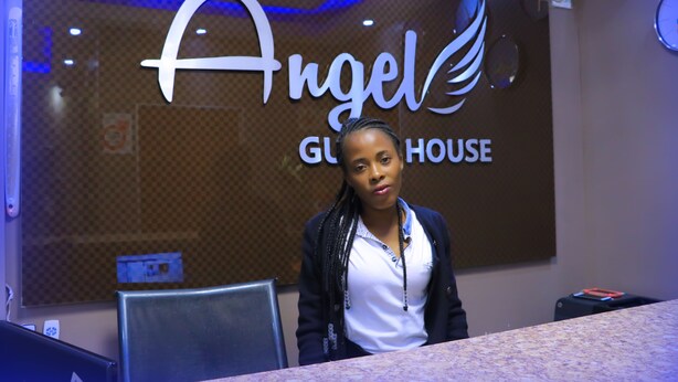 A person seated at a table in front of a wall with a sign that reads "Angel Glorious House." - Angel Guest House (Johannesburg)