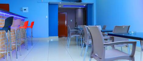 A modern dining area with a blue wall, a TV mounted above the bar, and a mix of metal and plastic chairs.