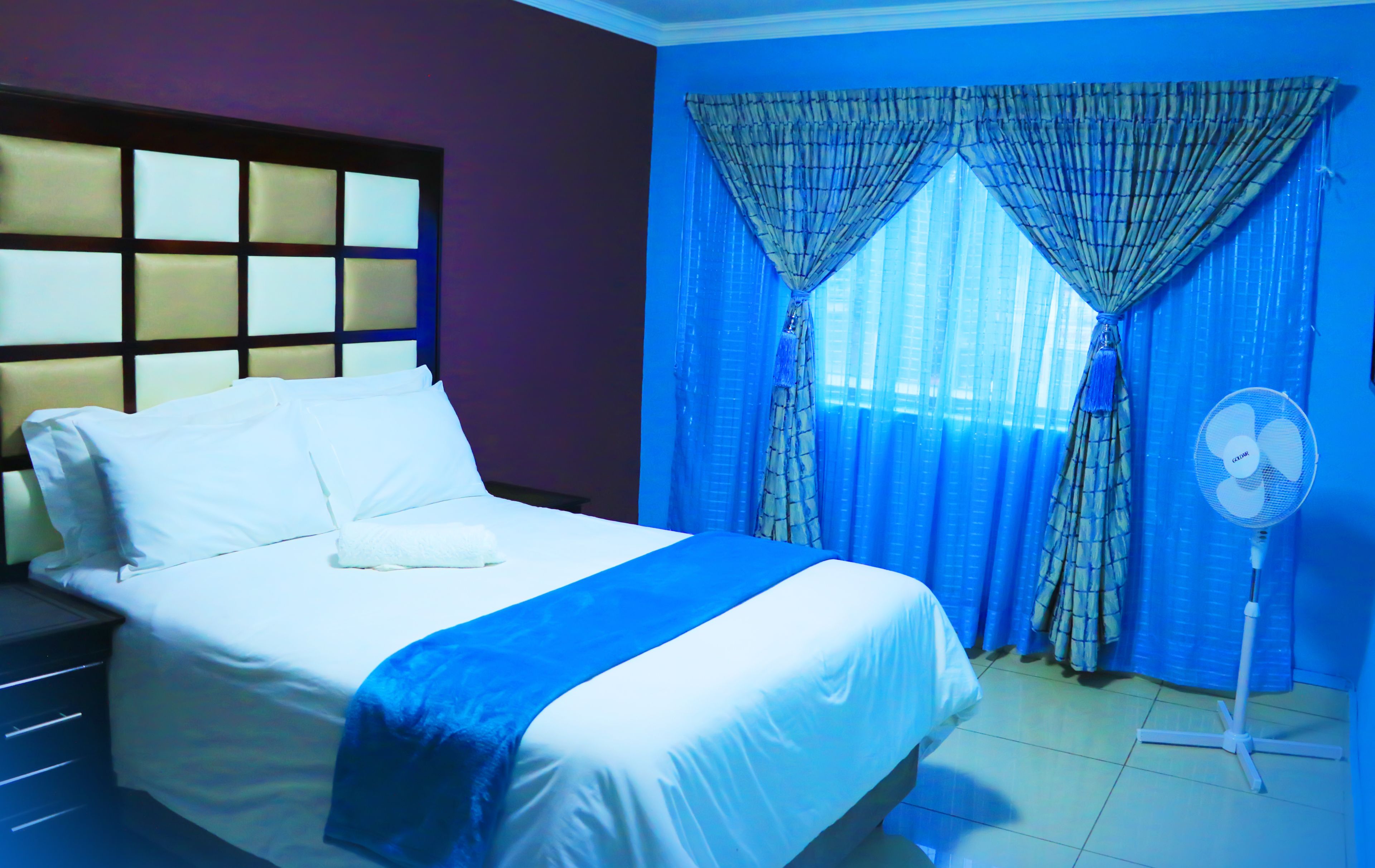 A neatly made bed with white linens and a blue accent, a standing fan, and a window with blue curtains.