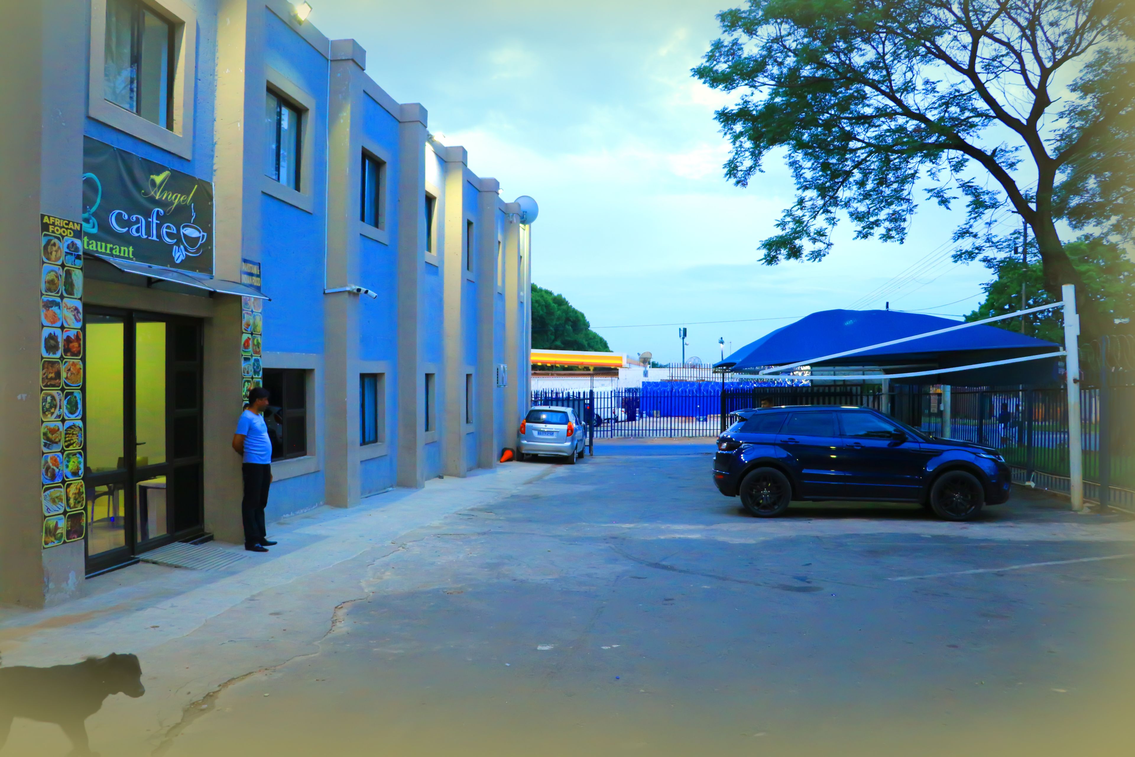 Vista da rua com um café chamado 'Angel Cafe' à esquerda, um carro azul estacionado à direita e uma estrutura de toldo azul ao fundo.
