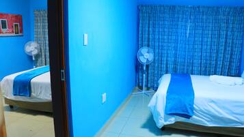 Habitació amb dos llits, un ventilador de peu i una finestra amb cortines blaves.