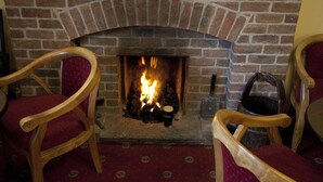 Une cheminée en briques avec un feu, entourée de chaises en bois avec des coussins rouges.