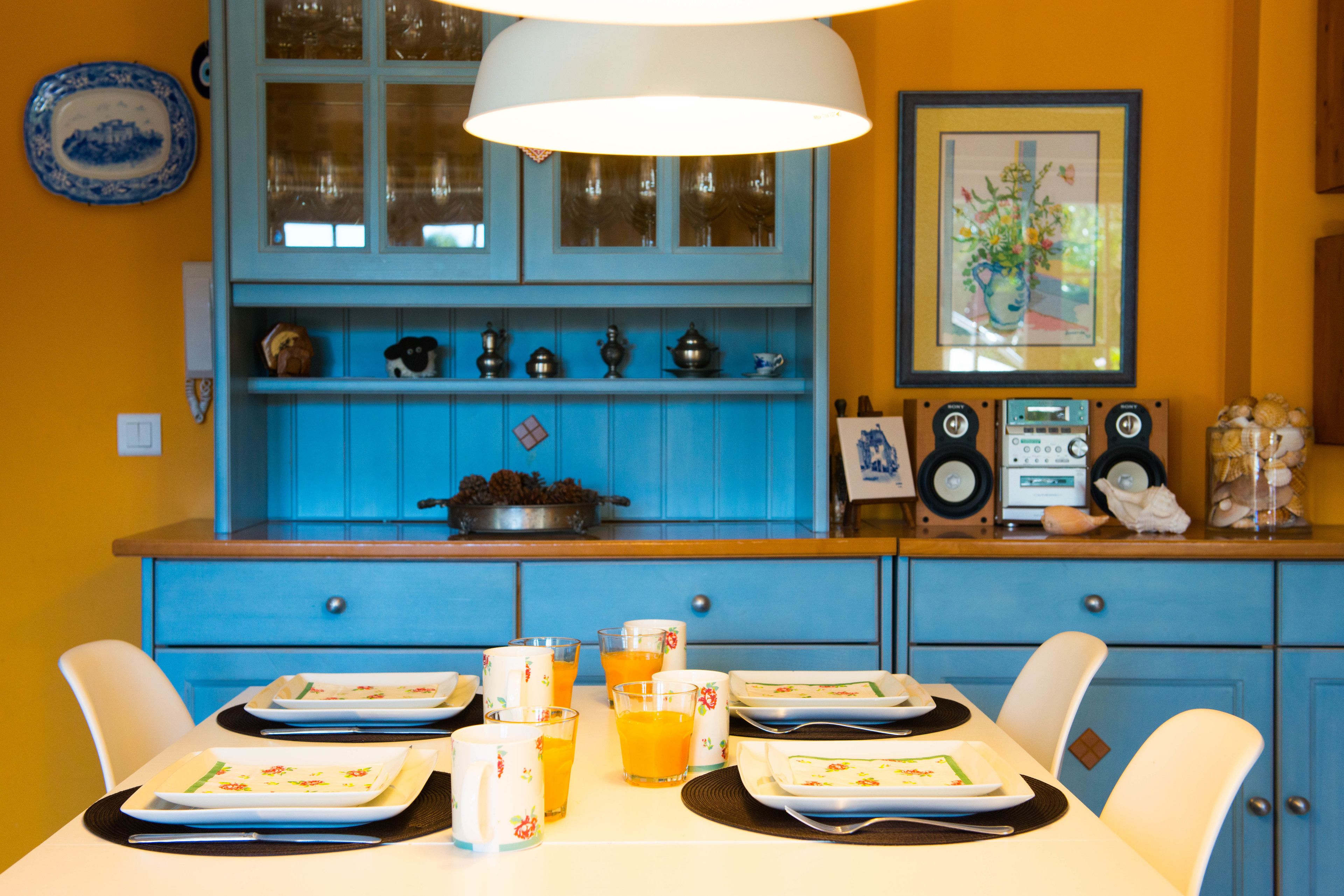 Un comedor con una mesa preparada para cuatro personas, un armario azul con puertas de cristal y un cuadro en la pared.