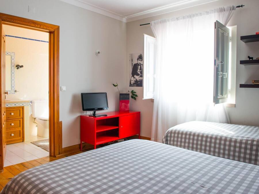 Quarto com duas camas, uma TV sobre um suporte vermelho, uma janela com cortinas e uma estante com livros.