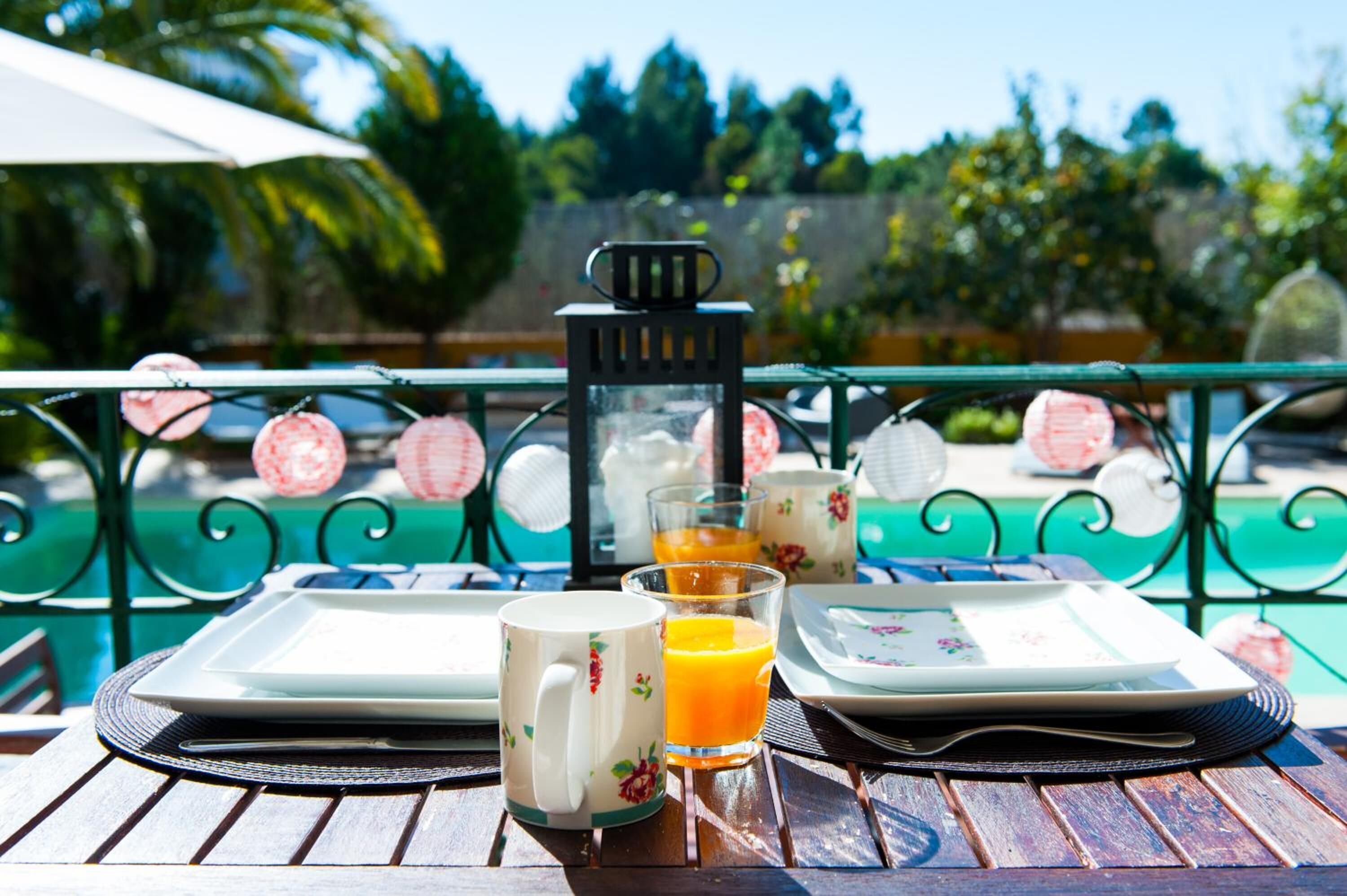 Desayuno con una mesa, platos, una taza y un vaso de zumo de naranja, con una piscina y faroles decorativos de fondo.