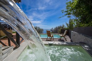En pool på taket med ett vattenfall, två solstolar i trä och utsikt över havet.