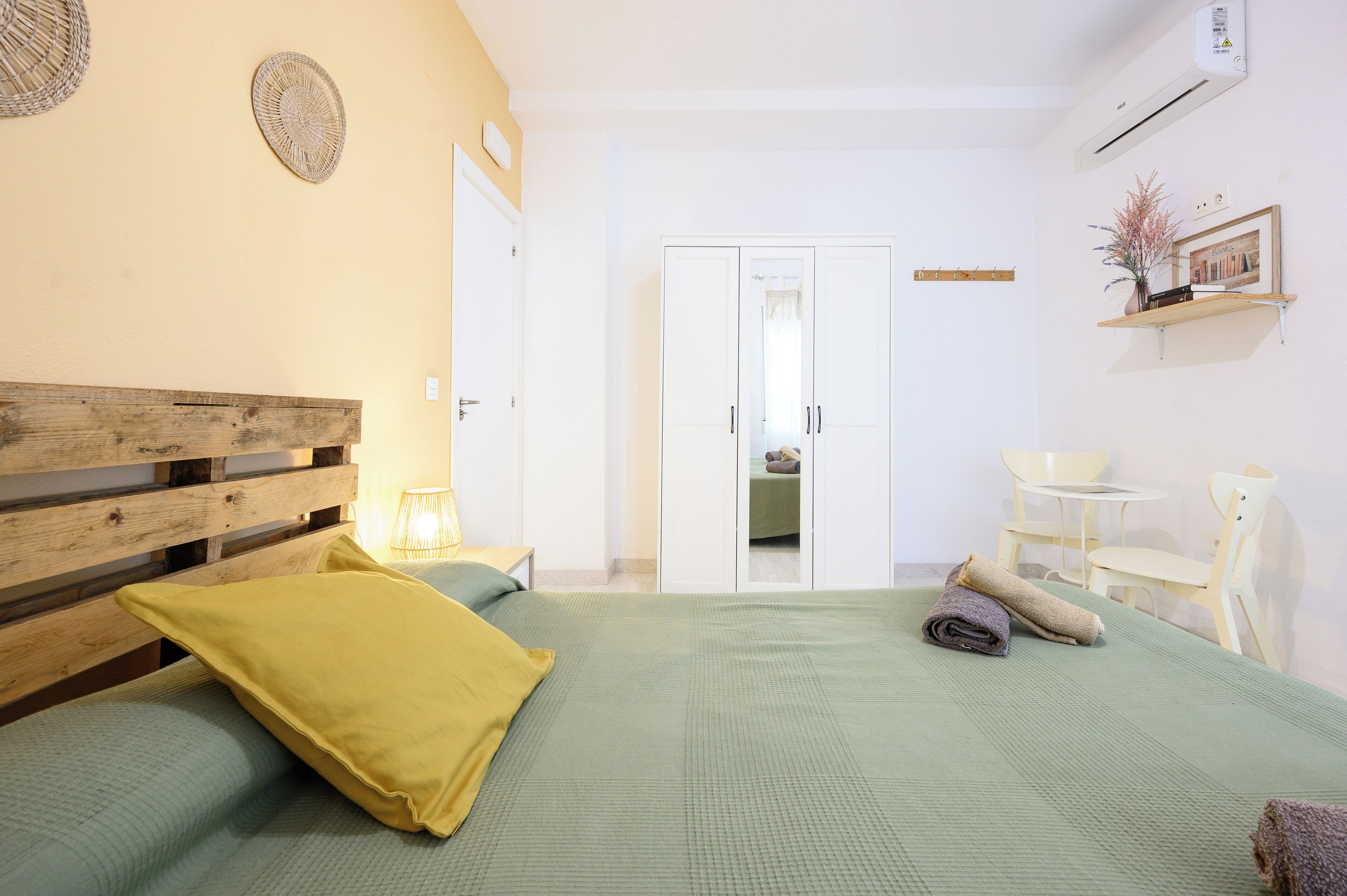 A bedroom with a wooden headboard, a green bedspread, and a yellow pillow. There is a white wardrobe, a small table with chairs, and a wall-mounted shelf with a plant.