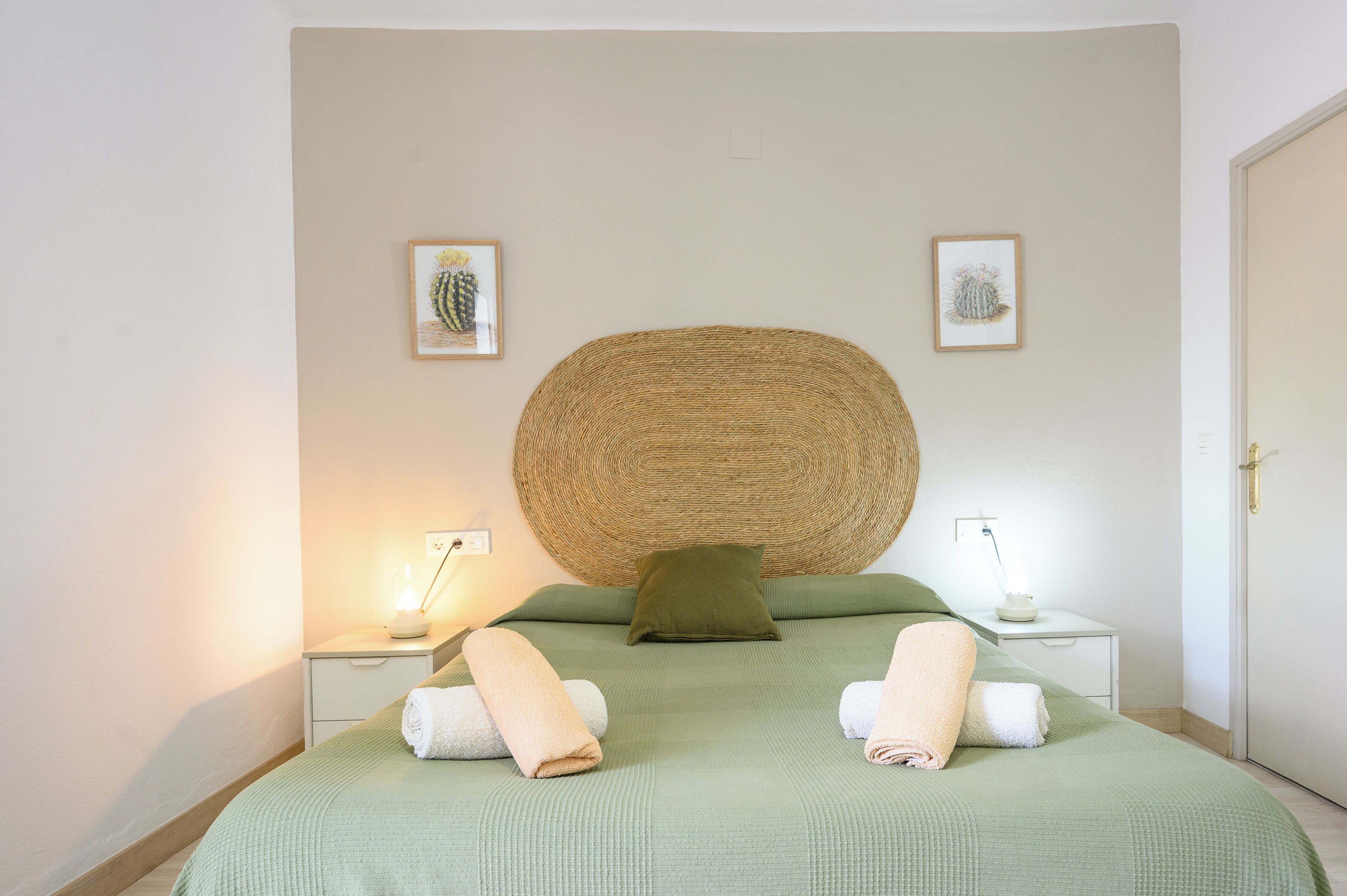 Una cama bien hecha con ropa de cama verde, dos mesitas de noche con lámparas, y dos obras de arte con motivos de cactus enmarcadas en la pared.