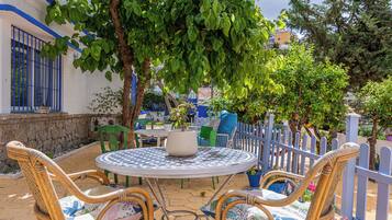 Un patio al aire libre con una mesa redonda y sillas de mimbre, rodeado por una cerca baja de color azul y abundante vegetación.