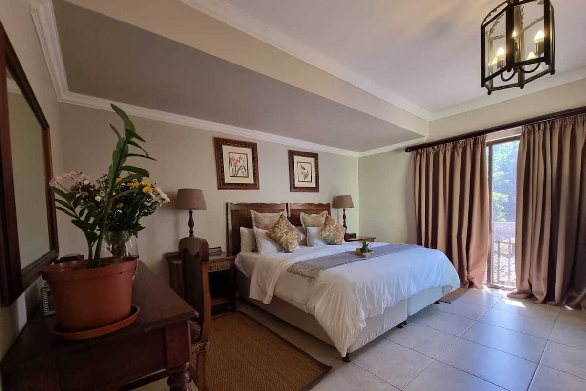 A hotel room with a large bed, two bedside tables with lamps, a potted plant, and a view of the outdoors through a window with curtains.