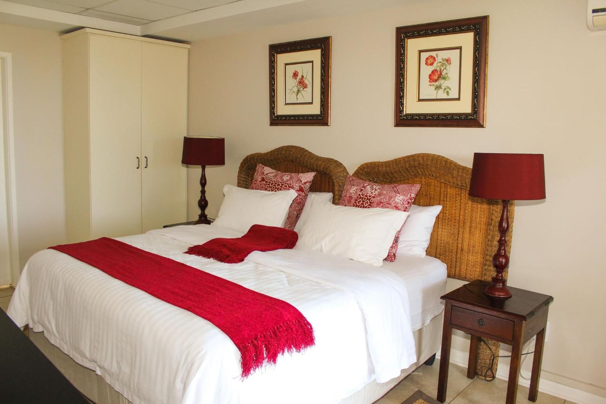 A neatly made bed with white and red bedding, two framed floral pictures on the wall, and a small bedside table with a lamp.