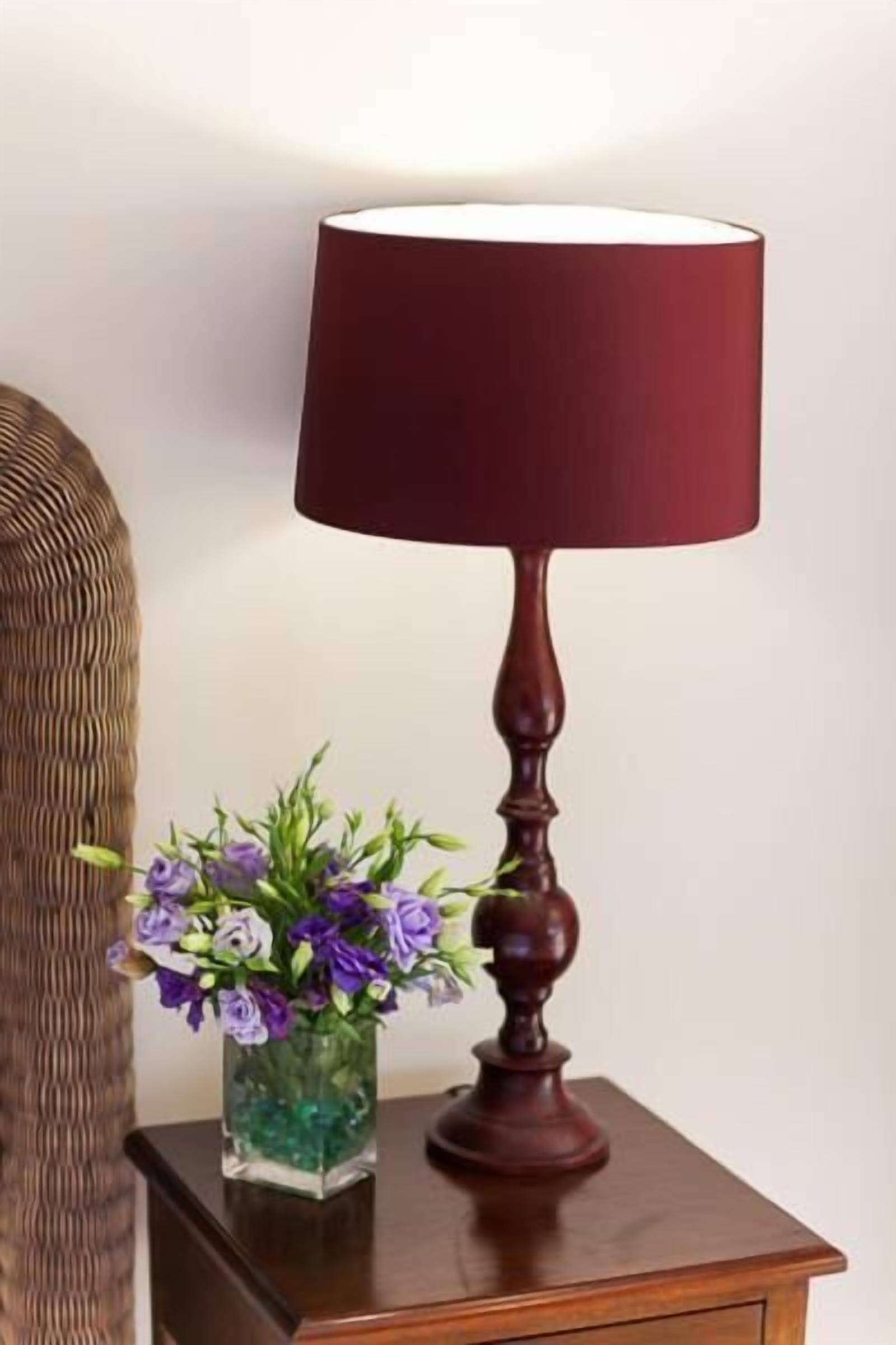 A wooden nightstand with a dark wooden lamp with a red shade and a clear vase with purple flowers.