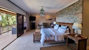 Une chambre spacieuse avec un grand lit, un mur en pierre, un ventilateur de plafond et un balcon offrant une vue sur les arbres.