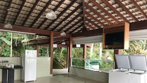 A covered outdoor kitchen area with a refrigerator, a sink, and a table. - Condomínio Residencial Grand Beach (Ubatuba)
