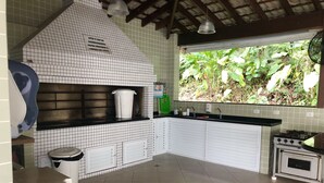 A kitchen with white cabinets, a stove, and a window overlooking greenery. - Condomínio Residencial Grand Beach (Ubatuba)
