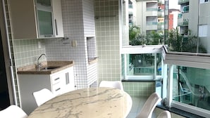 A modern kitchen with white cabinetry, a marble countertop, and a dining area with white chairs and a marble table. - Condomínio Residencial Grand Beach (Ubatuba)