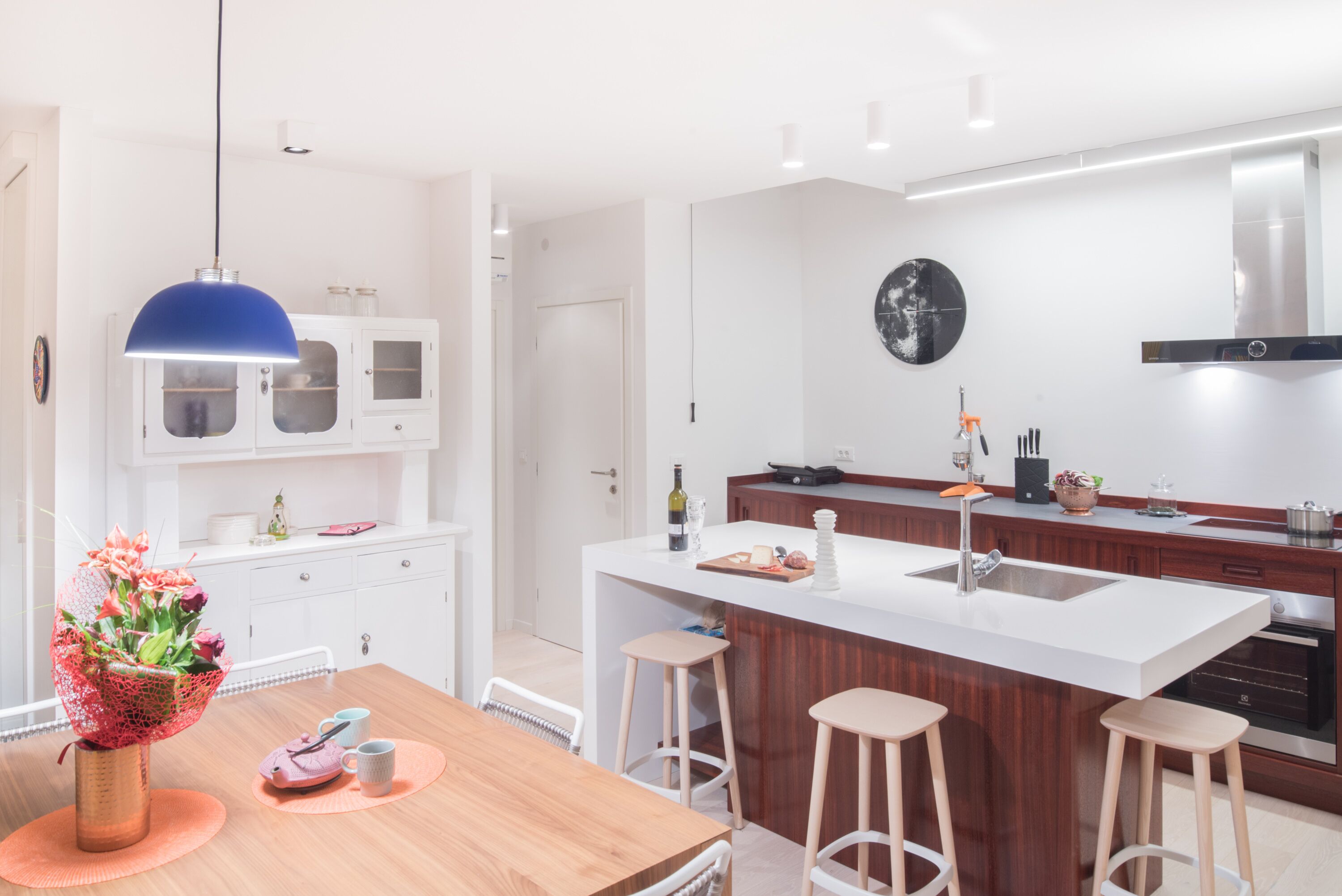 Een moderne keuken met witte kasten, een centraal eiland met krukken, een hanglamp en een decoratieve vaas met bloemen.