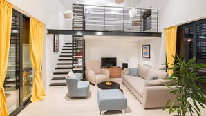 Ein modernes Wohnzimmer mit einer Couch, Sesseln, einem Couchtisch und einem Fernseher. Eine Treppe führt zu einer oberen Ebene.