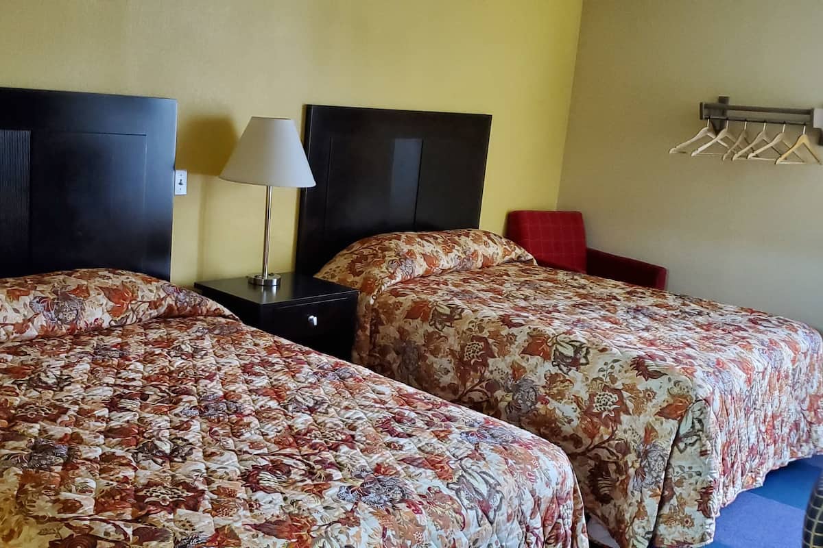 Ein Hotelzimmer mit zwei Betten, einem Nachttisch, einer Lampe, einem Fernseher und einem an der Wand befestigten Wäscheständer.