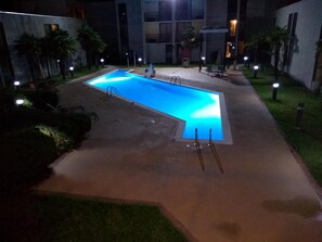 A swimming pool with a curved edge, surrounded by a concrete deck and small palm trees.