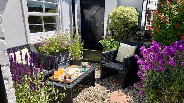 A patio area with outdoor seating, a small table with food and drinks, and various plants and flowers.