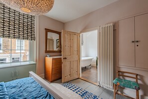 A bedroom with a bed, a wooden wardrobe, a dresser, a mirror, a radiator, and a bathroom visible through an open door.