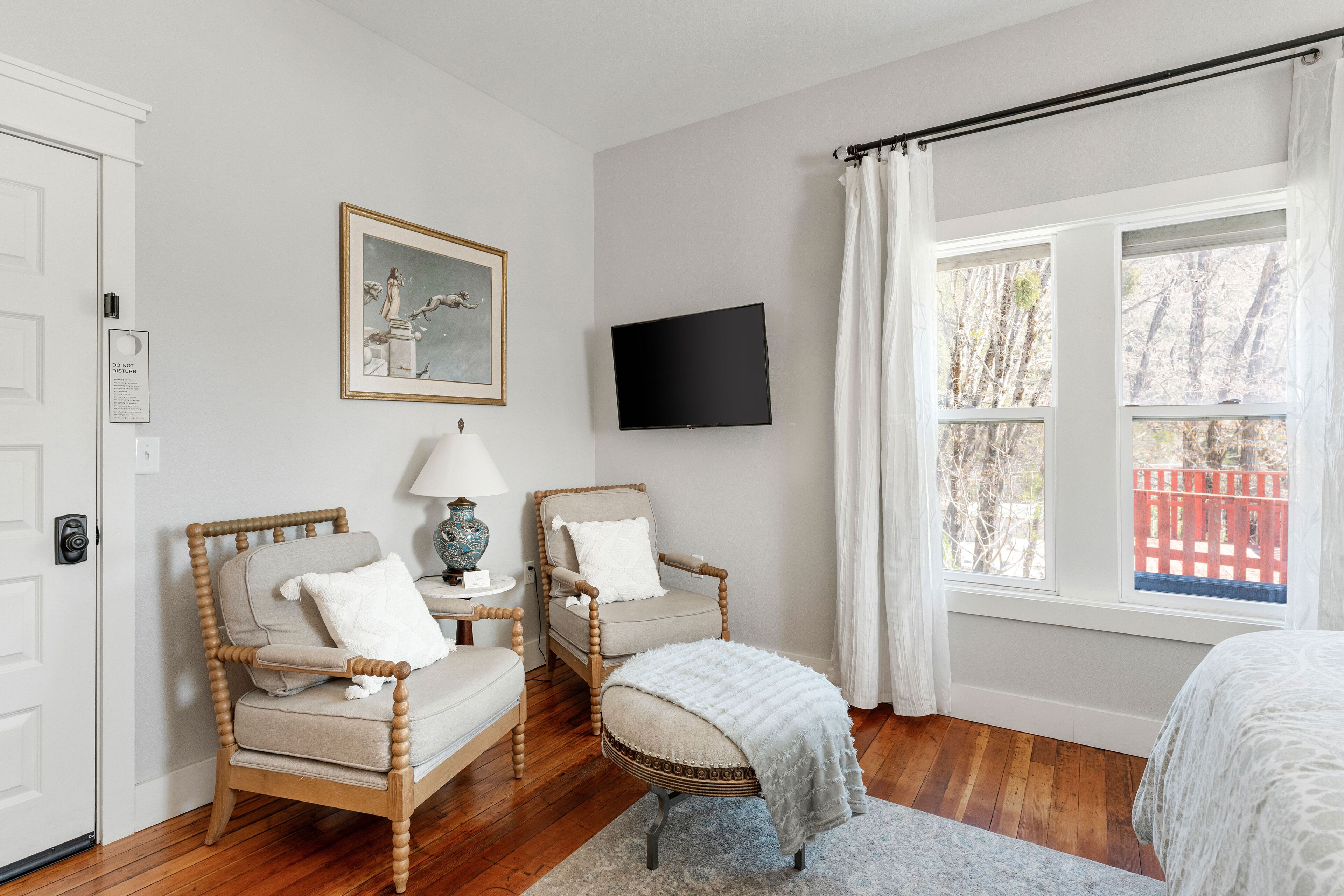A room with two wooden chairs, a television mounted on the wall, and a window with a view of trees and a red fence.