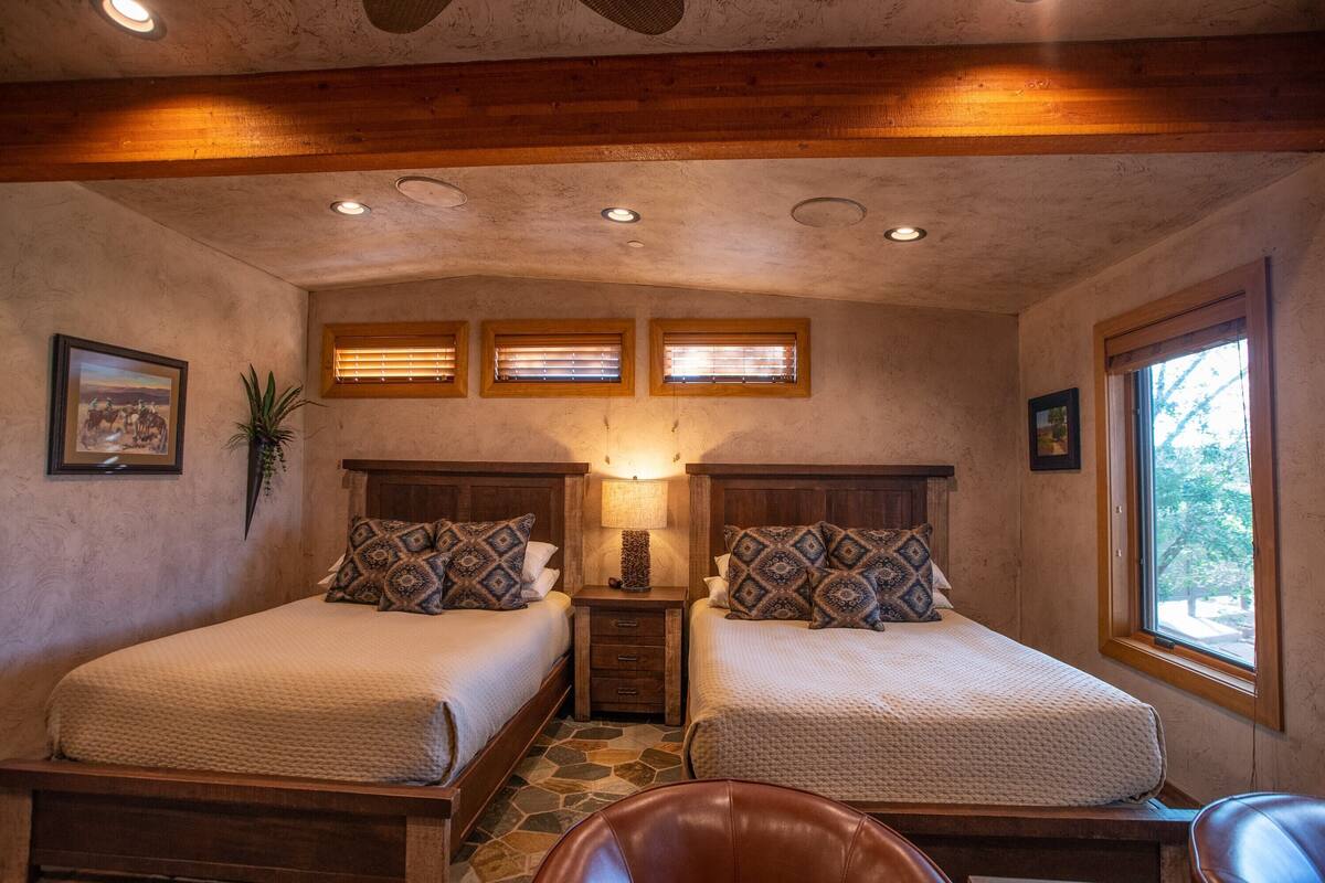A cozy bedroom with two beds, a nightstand, a lamp, and a window with blinds.