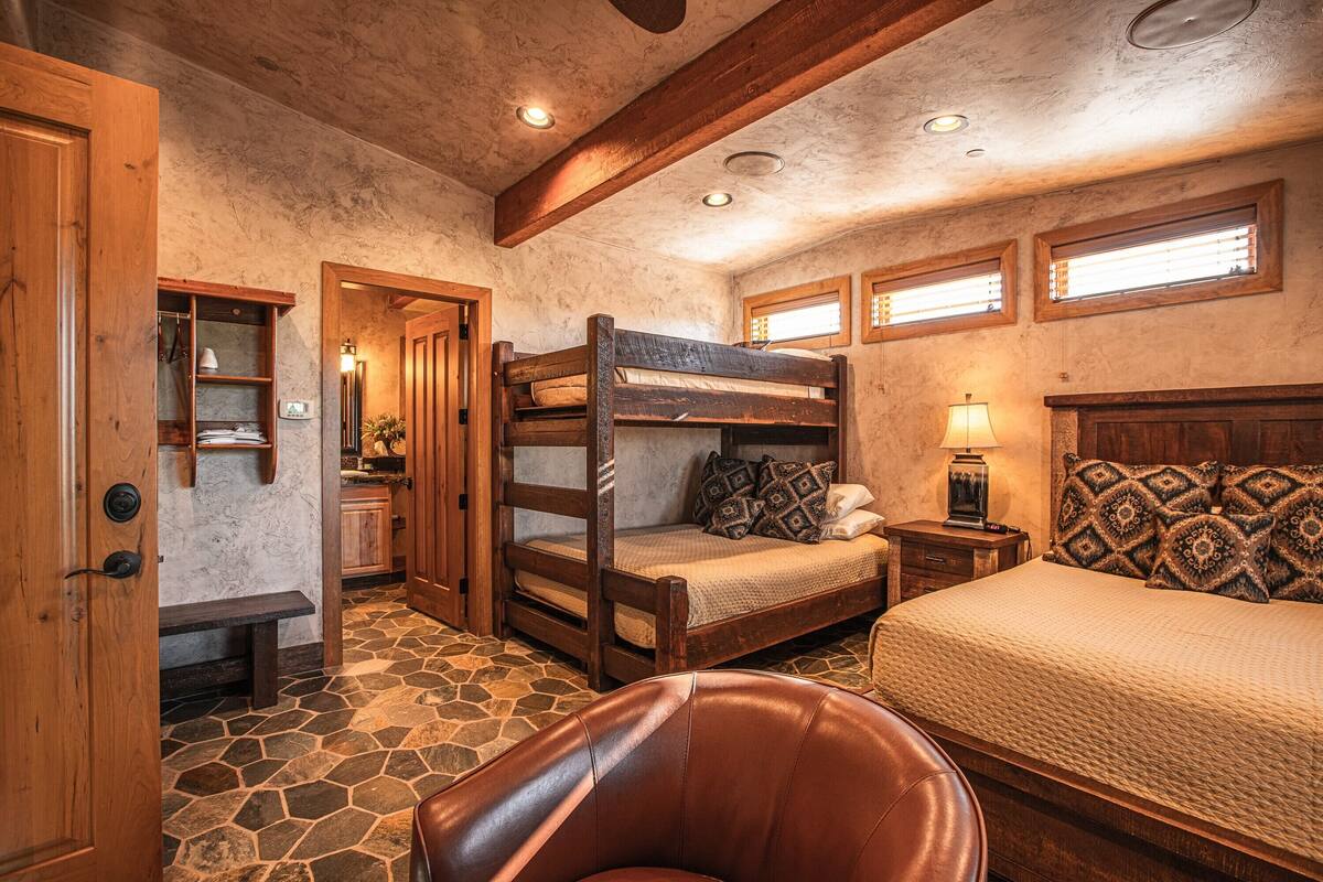 A rustic bedroom with a bunk bed, a single bed, a wooden bench, and a stone floor.