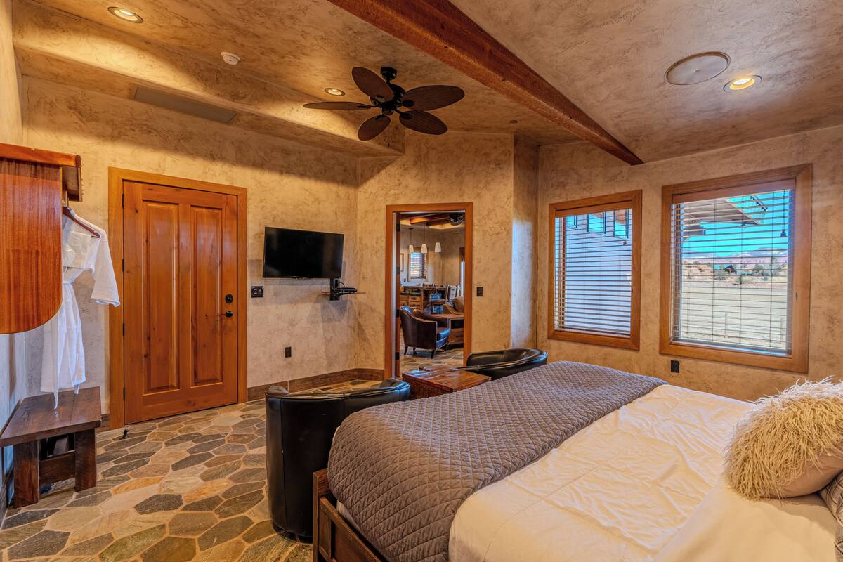 A bedroom with a stone floor, wooden ceiling beams, a bed with a quilted blanket, a ceiling fan, and a TV mounted on the wall.