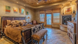 A bedroom with a bed, bench, dresser, fireplace, and artwork on the walls.