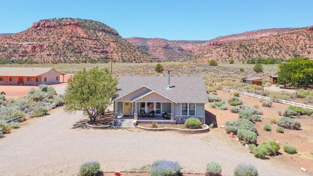 Uma casa térrea com varanda coberta e quintal cercado, rodeada por vegetação do deserto e colinas de rocha vermelha.