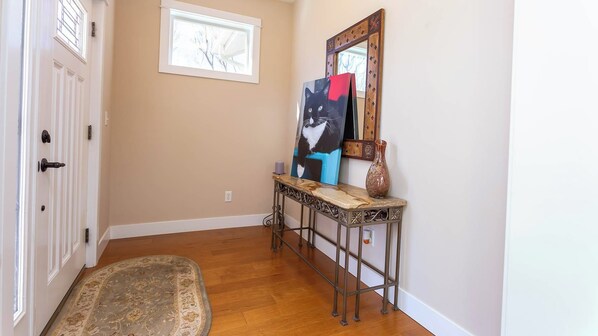 A hallway with a wooden floor, a rug, a console table with a vase, and a painting of a cat.